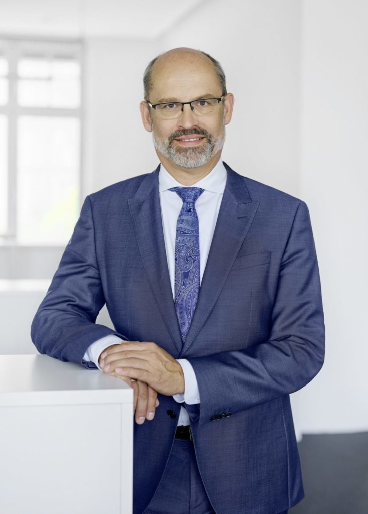 Halbtotale von Dr. Markus Zanner: Er steht in einem weißen Raum und lehnt sich an ein Sideboard.