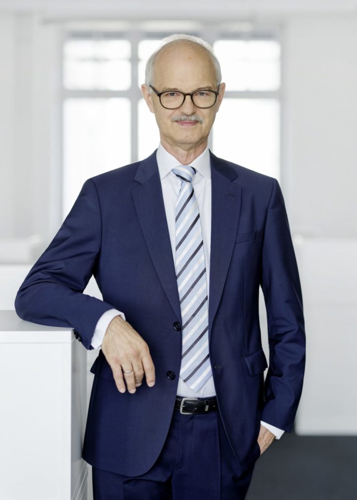 Halbtotale von Prof. Prömel. Er steht in einem weißen Raum und lehnt sich an ein Sideboard.