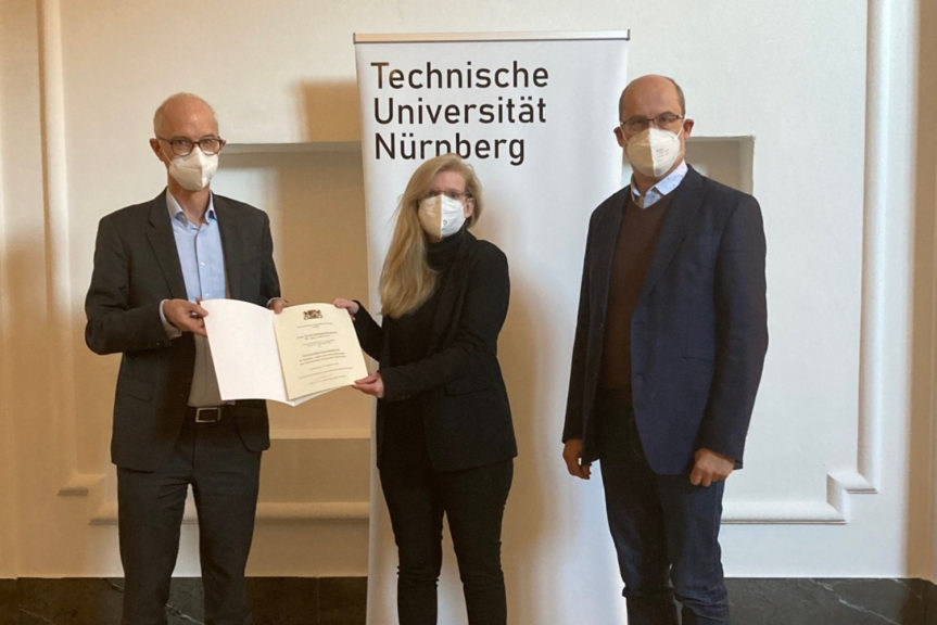 Prof. Prömel, Prof. Jahnke und Dr. Zanner stehen vor einem weißen Aufsteller mit dem schwarzen Schriftzug "Technische Universität Nürnberg". Prof. Prömel übergibt Prof. Jahnke die aufgeschlagene Urkunde.