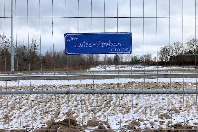 An einem Bauzaun hängt ein selbstgebasteltes Schild. Auf diesem steht "Dr,-Luise-Herzberg-Straße".