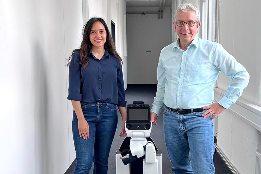 Reihaneh Mirjalili und Prof. Burgard stehen in einem Flur. Zwischen ihnen ist ein Pflegeroboter zu sehen.