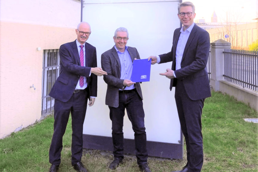Prof. Prömel und Markus Blume deuten auf eine blaue Mappe, die Prof. Burgard hält. Die drei stehen vor dem Eingang der TU Nürnberg.