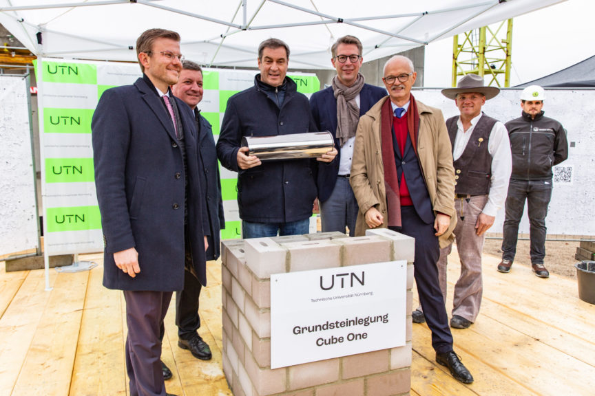 Die Redner stehen vor einem gemauerten Schach, auf dem "UTN Grundsteinlegung Cube One" steht. Dr. Söder hält in seinen Händen die Zeitkapsel.