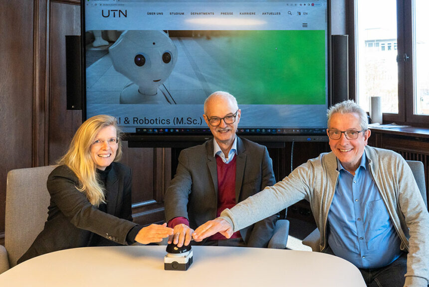 Prof. Jahnke, Prof. Prömel und Prof. Burgard sitzen vor einem großen Bildschirm. Auf diesem ist ein Roboter zu erkennen. Gemeinsam drücken sie einen Buzzer, der vor ihnen am Tisch steht.