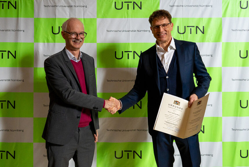 Prof. Prömel und Prof. Martin stehen vor einer grünen Pressewand mit dem Logo der UTN. Prof. Prömel schüttelt Prof. Martin die Hand. Dieser hält die aufgeschlagene Urkunde in die Kamera.