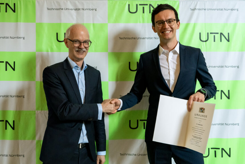 Prof. Prömel und Prof. Kipf stehen vor einer grünen Pressewand mit dem Logo der UTN und schütteln sich die Hand. Prof. Kipf hält die Urkunde in die Kamera.
