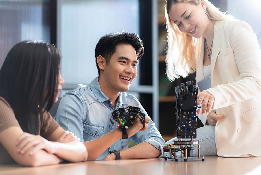 Drei Personen experimentieren mit einem Roboterhandschuh. Der Mann in der Mitte trägt diesen, die Frau rechts deutet auf das Exponat. Eine weitere Frau auf der linken Seite verfolgt das Experiment.