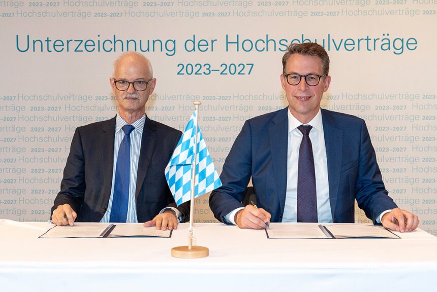 Prof. Prömel und Markus Blume sitzen vor zwei aufgeschlagenen Mappen. Auf der Wand hinter ihnen steht "Unterzeichnung der Hochschulverträge 2023-2027". Auf dem Tisch steht eine Bayerische Fahne.