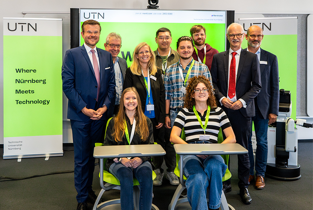 Die Studierenden stehen und sitzen gemeinsam mit dem Präsidium und OB Marcus König vor einem grünen Screen. Auf der linken Seite ist ein Roll-up mit der Aufschrift "Where Nuremberg meets Technology" zu sehen.