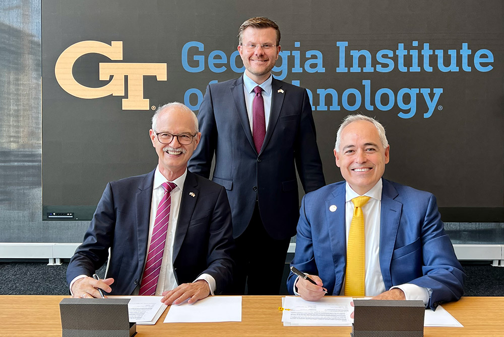 Prof. Prömel und Prof. Cabrera sitzen an einem Tisch vor einem Banner mit der Aufschrift "Georgia Institute of Technology". Auf dem Tisch liegen die aufgeschlagenen Vertragsdokumente, Hinter den beiden steht OB Marcus König.