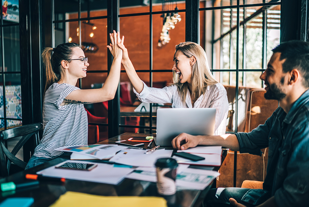 Zwei Frauen sitzen an einem Tisch, auf dem Dokumente ausgebreitet sind, und geben sich ein High-Five.