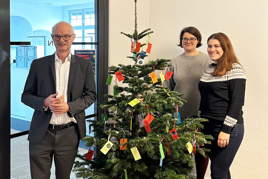 Ein Mann und zwei Frauen stehen um einen Weihnachtsbaum, an dem bunte Zettel hängen.