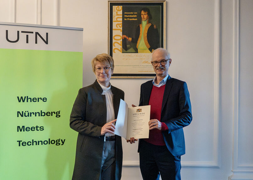 Prof. Prömel und Prof. Grimm stehen neben einem grünen Rollup mit dem Logo der UTN. Prof. Prömel übergibt Prof. Grimm die Urkunde.