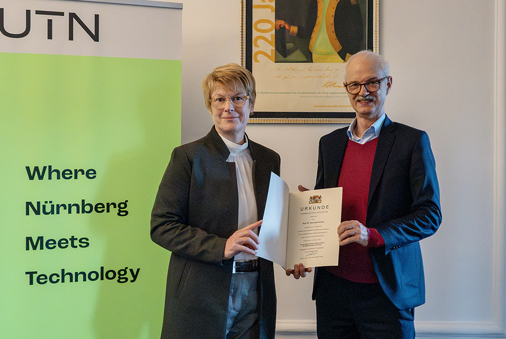 Prof. Prömel und Prof. Grimm stehen neben einem grünen Rollup mit dem Logo der UTN. Prof. Prömel übergibt Prof. Grimm die Urkunde.