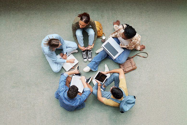 Six people sit on the floor in a circle, engaging with laptops, notebooks, and a tablet.