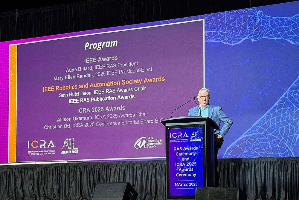 Professor Wolfram Burgard presenting the program items for the IEEE RAS Awards Ceremony at ICRA 2025 in Atlanta, stage with conference banner in the background.