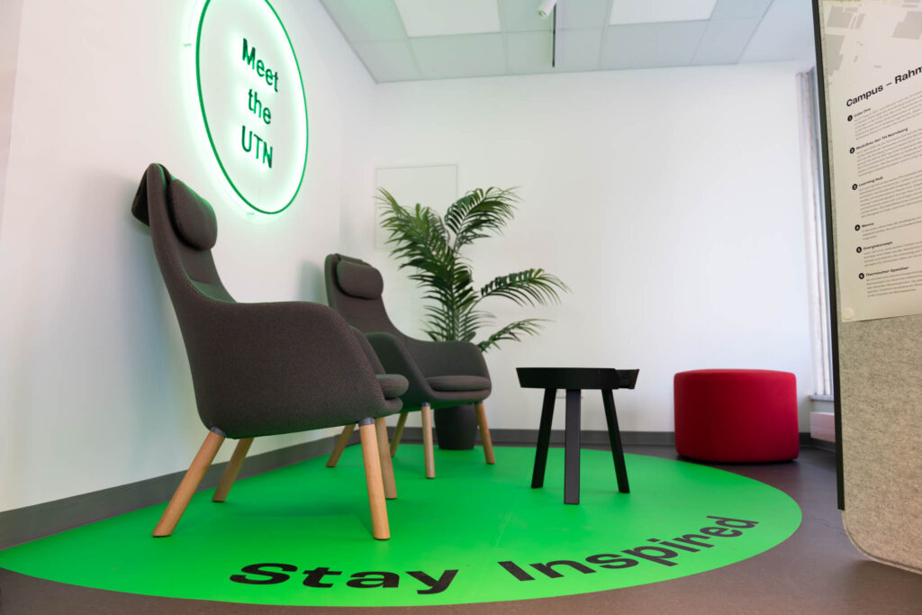 Zwei graue Sessel in der UTN Science Lounge. Sie stehen auf einer grünen Fläche mit der Aufschrift "Stay Inspired".