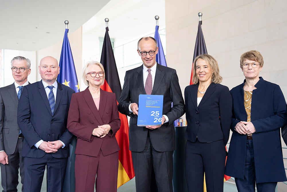 Bundeskanzler Friedrich Merz mit fünf Mitgliedern des Sachverständigenrats (Wirtschaftsweise); der Kanzler hält das Jahresgutachten 2025/26. Im Hintergrund EU- und Deutschlandflaggen.