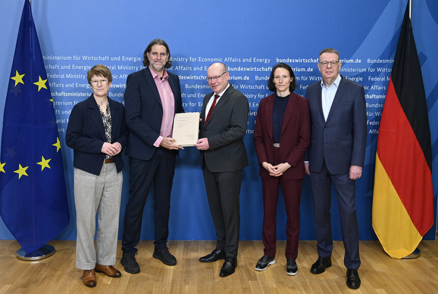 Fünf Personen stehen vor einer blauen Wand mit der Aufschrift „Bundesministerium für Wirtschaft und Energie“, links die EU-Flagge und rechts die Deutschlandflagge; in der Mitte überreichen zwei Personen ein Gutachten/Dokument, die anderen stehen daneben und schauen in die Kamera.