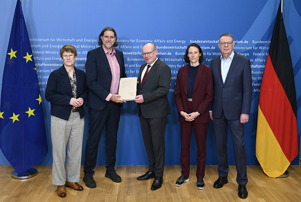 Fünf Personen stehen vor einer blauen Wand mit der Aufschrift „Bundesministerium für Wirtschaft und Energie“, links die EU-Flagge und rechts die Deutschlandflagge; in der Mitte überreichen zwei Personen ein Gutachten/Dokument, die anderen stehen daneben und schauen in die Kamera.
