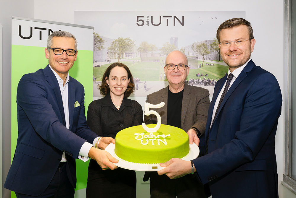 Vier Personen beim feierlichen Tortenanschnitt zum UTN-Jubiläum: grüne Torte mit großer ‚5‘ vor UTN-Hintergrund