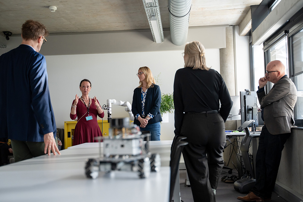 Eine Person erklärt einer kleinen Gruppe in einem Technikraum etwas; im Vordergrund steht ein unscharfer mobiler Roboter auf einem Tisch.