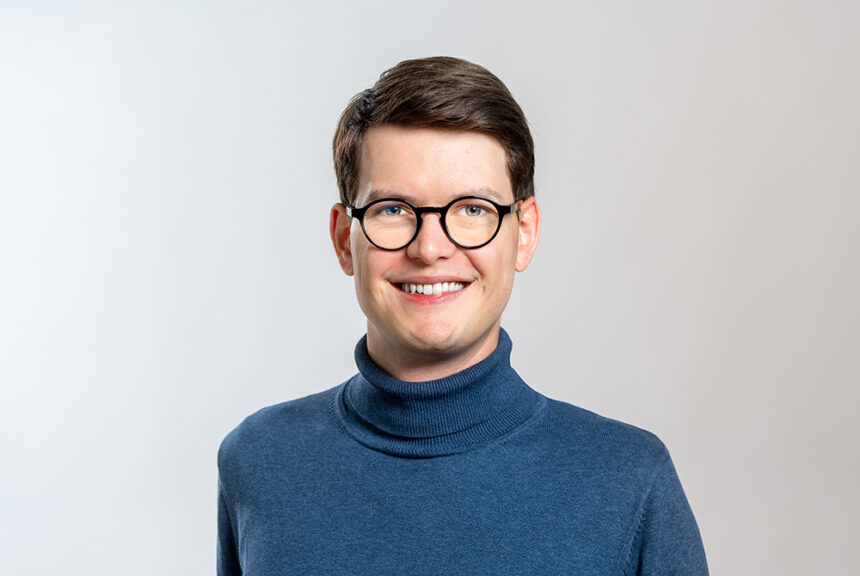 Porträt von Prof. Dr. Vincent Fortuin, Probabilistic Machine Learning, mit Brille und blauem Rollkragenpullover vor hellem Hintergrund.