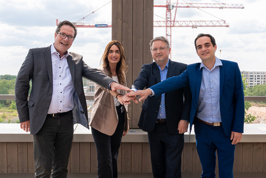 Peter Will (EnergieRegion), Arina Zonner (UTN), Dr. Jens Hauch (EnergieRegion) und Dr. Christian Sölch (UTN) legen gemeinsam ihre Hände übereinander. Im Hintergrund ist die Baustelle des UTN-Campus mit mehreren Baukränen zu sehen.